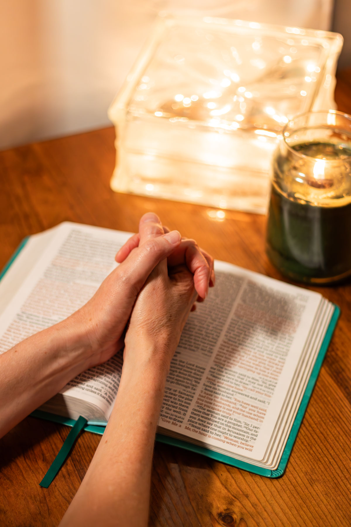 praying hands on an open Bible with a candle in the background.